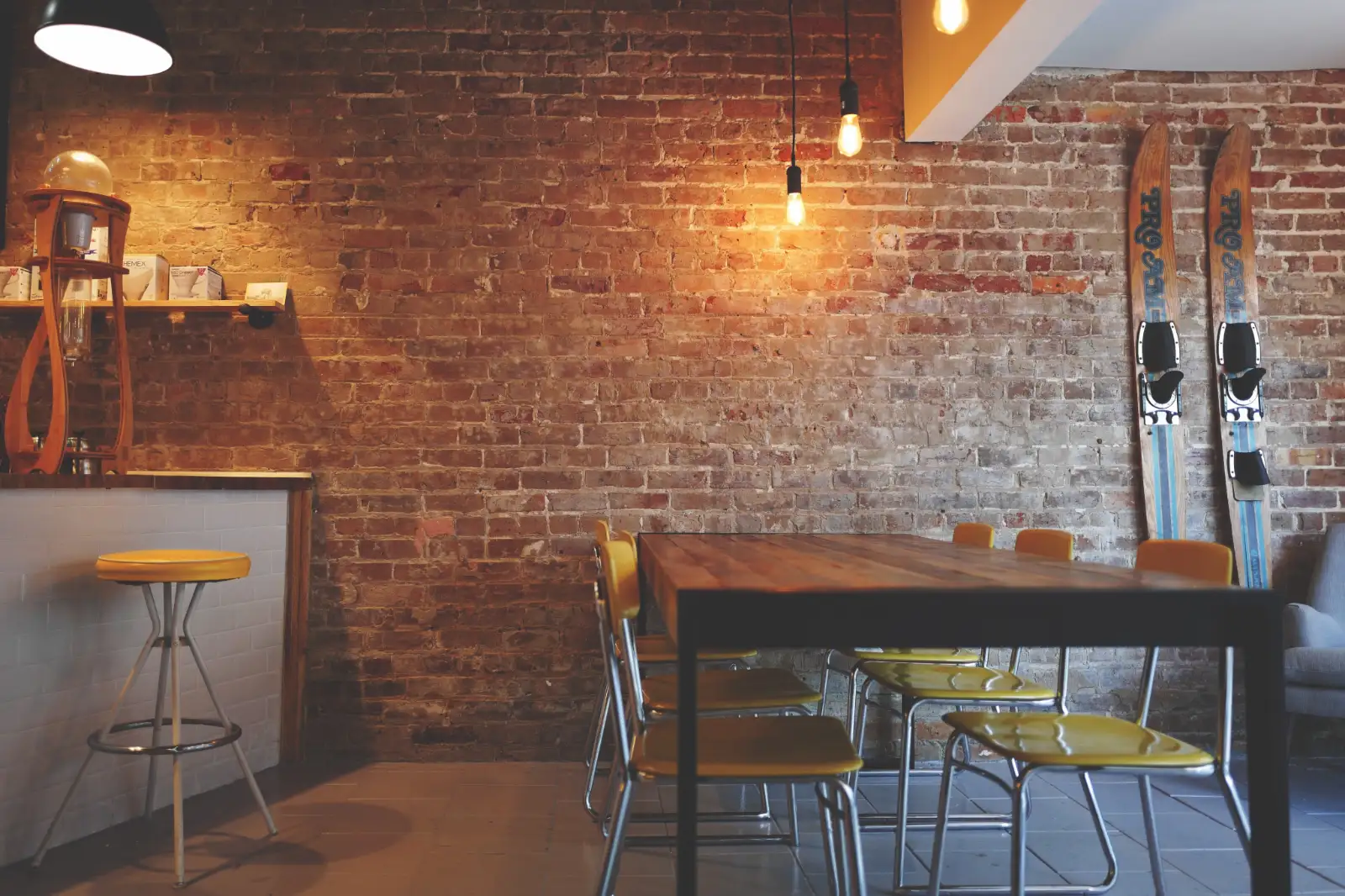 A coffee shop corner with a brick wall, a long wooden table and yellow chairs