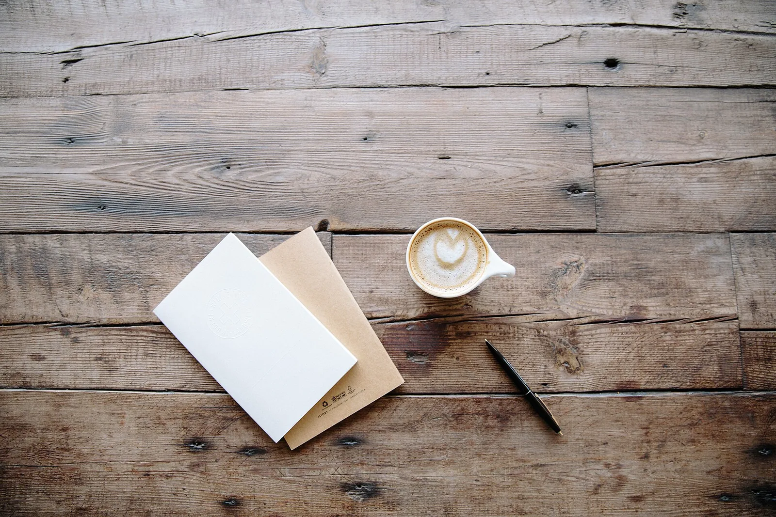 A cup of coffee beside two notebooks and a pen on a wooden table